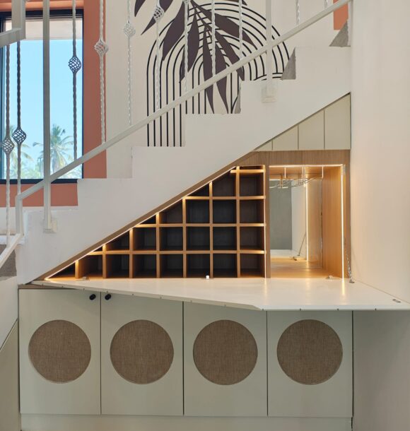 Under-stair bar unit with backlit wooden bottle shelves, cream lower cabinets, circular cane shutter details, fold-out white counter, staircase railing, and decorative wall art.