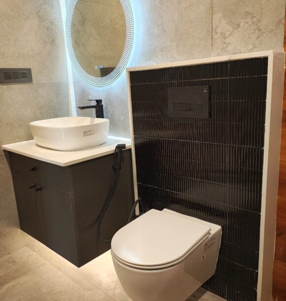 Modern bathroom with round backlit mirror, white vessel basin, black vanity cabinet, wall-mounted toilet, black textured tile partition, grey stone wall tiles, and warm floor lighting.