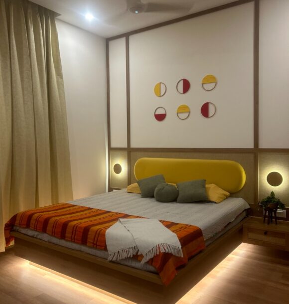 Modern bedroom with yellow upholstered headboard, wooden wall panelling, circular wall decor, bedside lights, beige curtains, wooden flooring, and warm under-bed lighting.