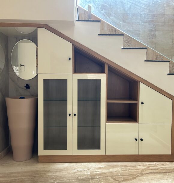 Under-stair storage unit with cream cabinets, wooden framing, fluted glass doors, open shelves, black handles, compact wash basin, round mirror, and glass staircase railing.