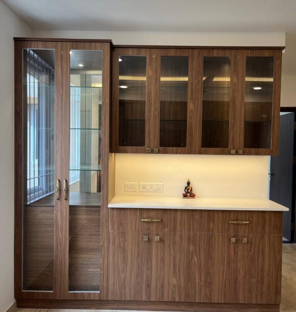 Wooden crockery unit with glass display cabinets, tall side storage, white countertop, warm under-cabinet lighting, brass hardware, and marble flooring.