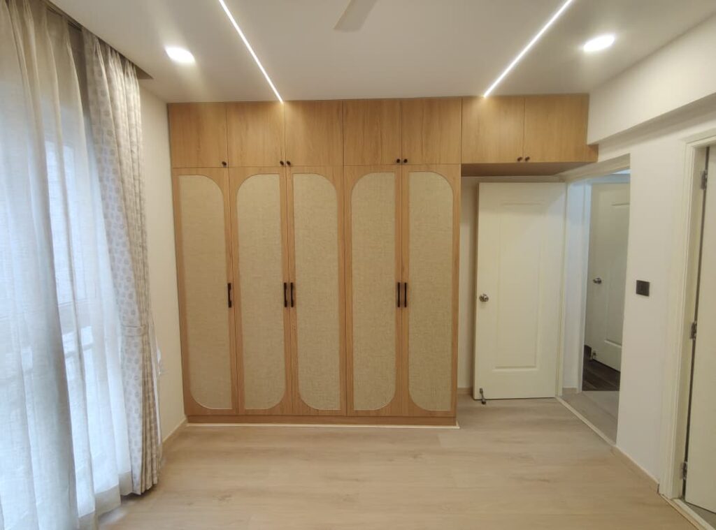 Bedroom with double wooden wardrobe, overhead loft cabinets, arched fabric panel shutters, black handles, side mirror, light flooring, curtains, and linear ceiling lights.