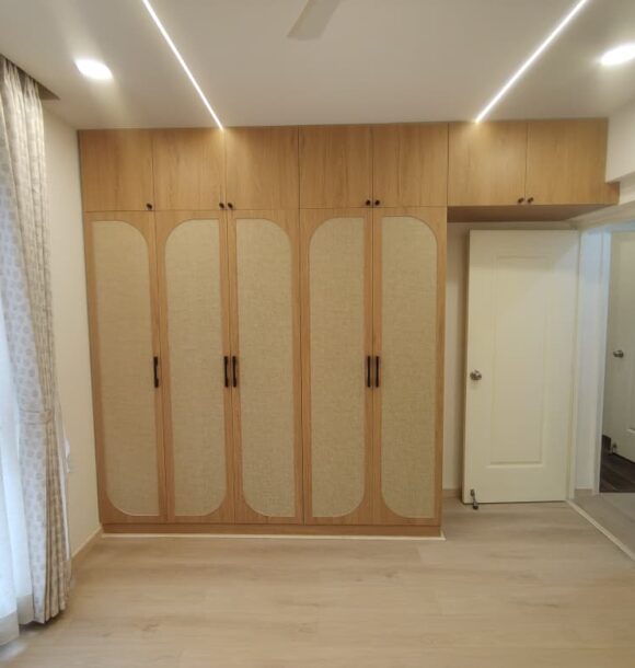 Bedroom with double wooden wardrobe, overhead loft cabinets, arched fabric panel shutters, black handles, side mirror, light flooring, curtains, and linear ceiling lights.
