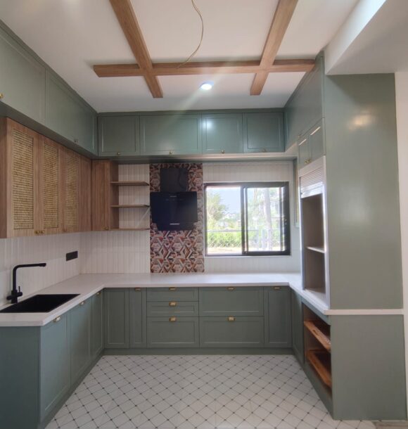 Modern kitchen with sage green cabinets, wooden ceiling beams, cane cabinet shutters, white countertops, patterned backsplash tiles, black sink, brass handles, and large window.