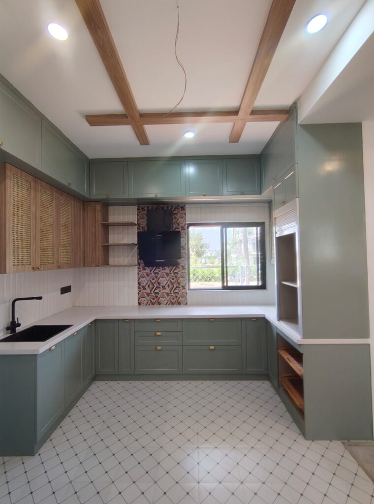 Modern kitchen with sage green cabinets, wooden ceiling beams, cane cabinet shutters, white countertops, patterned backsplash tiles, black sink, brass handles, and large window.