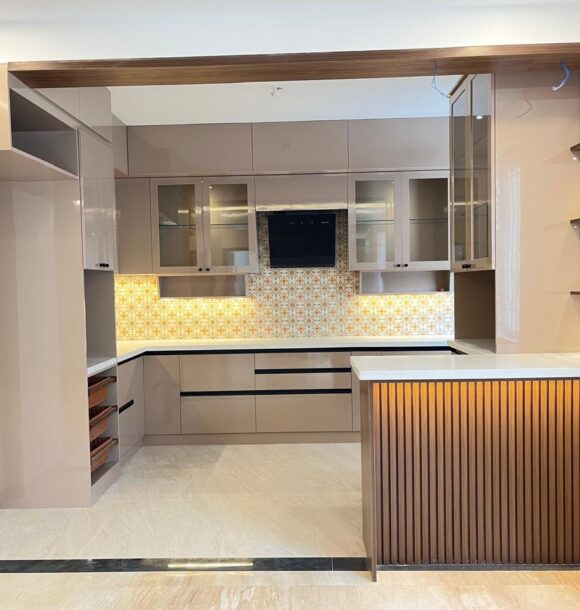 Modern kitchen with glossy taupe cabinets, patterned backsplash, glass-front wall cabinets, white countertops, wooden ceiling frame, open shelves, and fluted wooden breakfast counter.