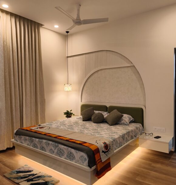 Modern master bedroom with curved feature wall, upholstered headboard, neutral bedding, pendant light, beige curtains, wooden flooring, and warm under-bed lighting.