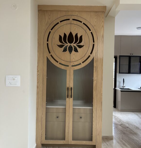Wooden puja unit with lotus motif cutwork, circular design detail, fluted glass cabinet shutters, black handles, and warm natural wood finish.