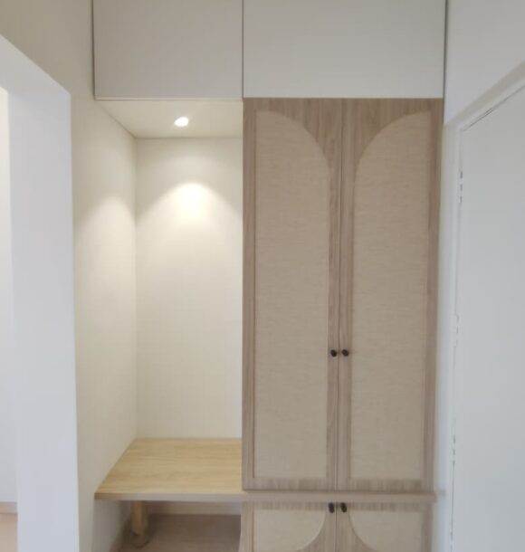 Compact single wardrobe with light wooden finish, arched panel shutters, overhead white cabinets, small black handles, built-in side bench, and recessed ceiling light.