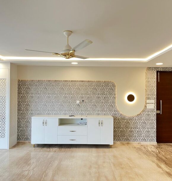 Living room wall with white TV console, integrated puja unit with decorative cutwork doors, patterned wallpaper, wooden main door, wall light, cove ceiling lighting, and marble flooring.