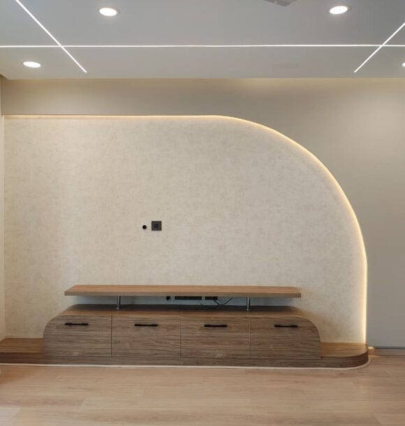 Modern living room TV unit with curved textured feature wall, warm backlighting, wooden storage console, rounded cabinet edges, beige curtains, linear ceiling lights, and wooden flooring.