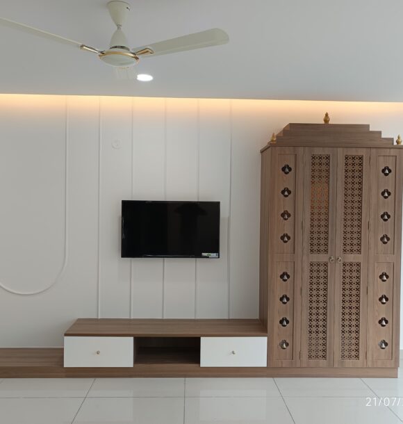 Real Photo Living room TV unit with white panelled wall, low wooden console, warm cove lighting, and traditional wooden puja unit with decorative cutwork doors and bell details.
