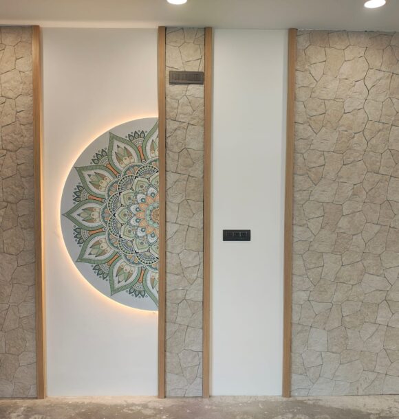 Decorative interior wall with stone-textured panels, vertical wooden trims, white wall sections, illuminated mandala artwork, recessed ceiling lights, and warm accent detailing.