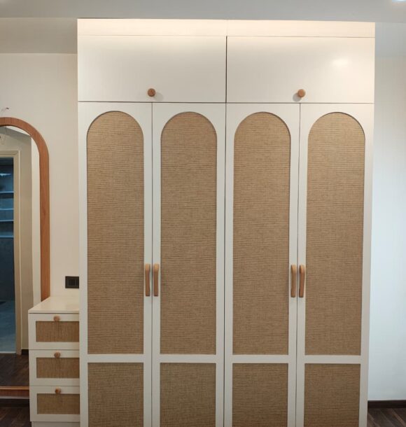 White bedroom wardrobe with arched cane panel shutters, wooden handles, overhead cabinets, matching dressing table, curved mirror frame, drawers, and dark wooden flooring.
