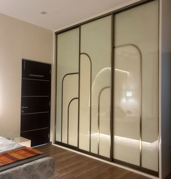 Modern bedroom with large sliding wardrobe, glossy cream glass shutters, dark metal framing, curved line detailing, wooden flooring, black door, and partial bed view.