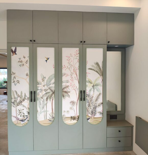 Sage green bedroom wardrobe with botanical printed shutters, bird and plant motifs, rounded panel detailing, black handles, overhead cabinets, and built-in dressing table.