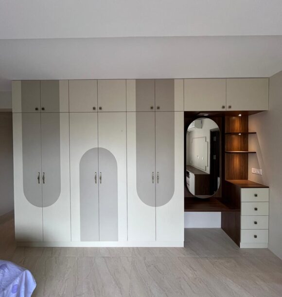 Modern bedroom wardrobe with white and grey shutters, arched panel detailing, overhead loft cabinets, integrated wooden dressing table, oval mirror, open shelves, drawers, and marble flooring.