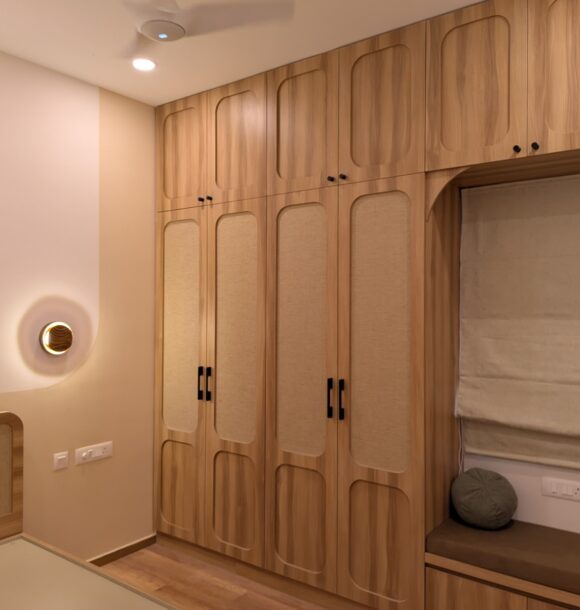Wooden bedroom wardrobe with rounded panel design, fabric inserts, black handles, integrated overhead cabinets, wall light, Roman blind, and cushioned window seating.
