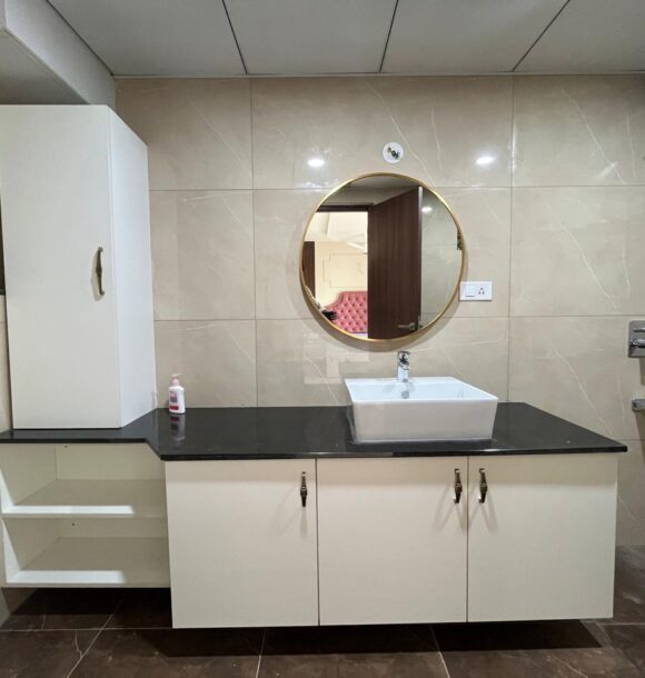 Modern washroom vanity with white storage cabinet, black countertop, rectangular vessel basin, round gold-rimmed mirror, tall side cabinet, open shelves, beige wall tiles, and shower area.