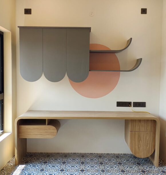 Modern workstation with light wooden desk, grey wall cabinets, curved shelves, peach circular wall accent, patterned floor tiles, and large windows on both sides.